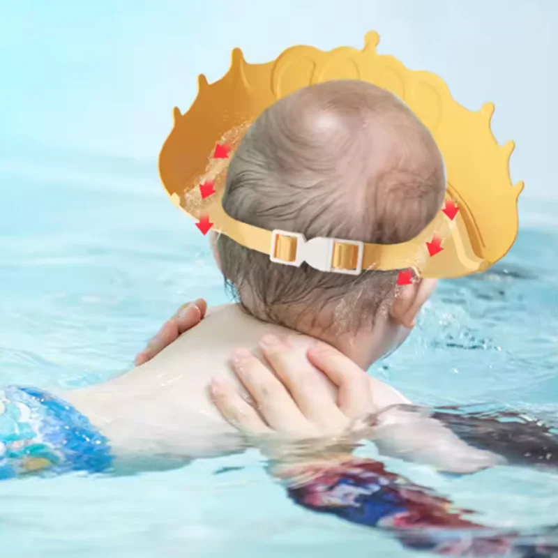 A baby is shown wearing a yellow bath shield cap with ear protection while in water.