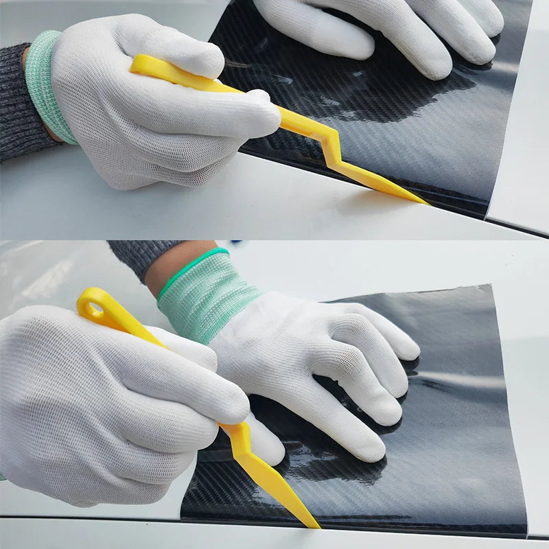 A hand wearing a white glove uses a yellow vinyl wrap tool on a dark surface.