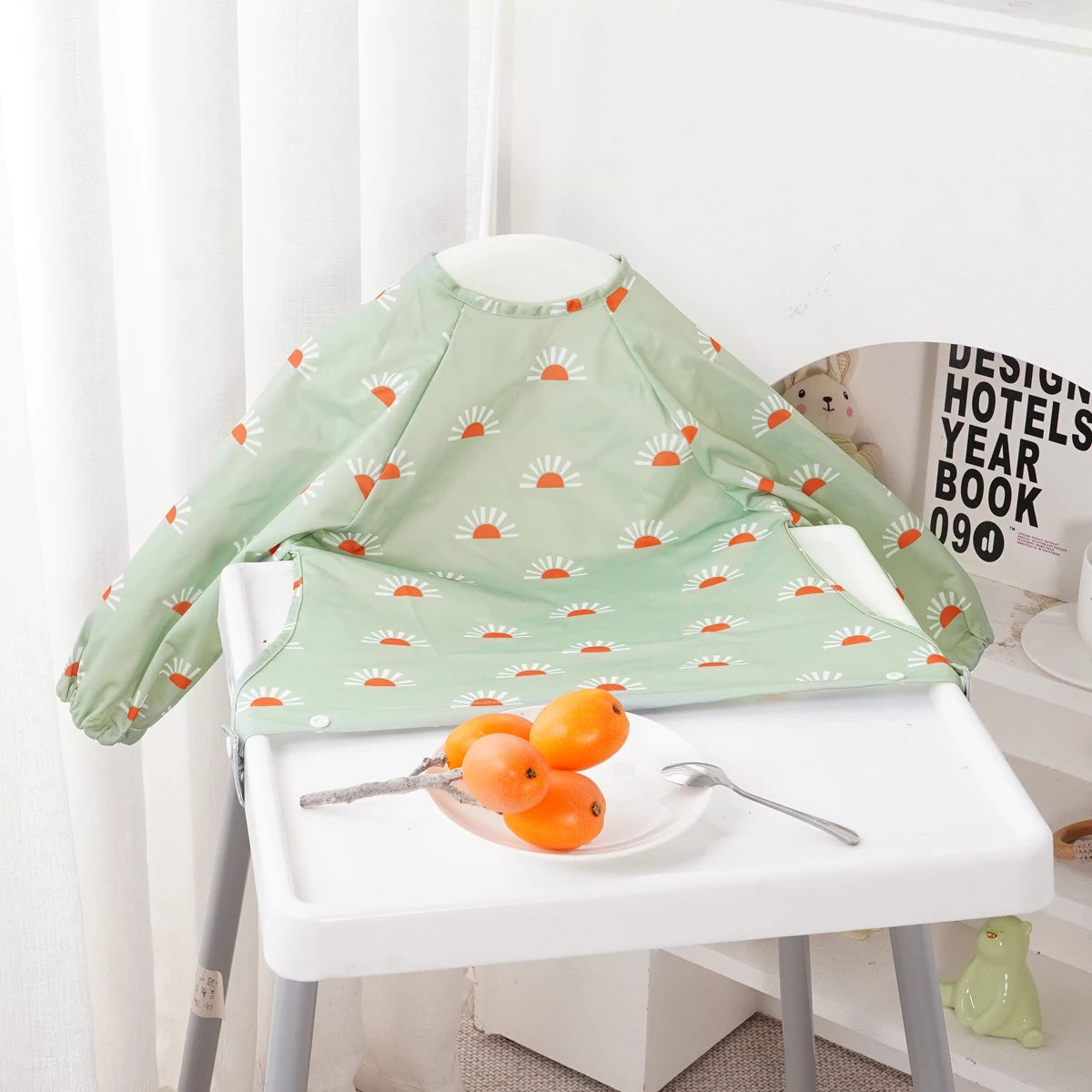A light green long-sleeve waterproof baby bib with a repeating orange flower pattern displayed on a white table.