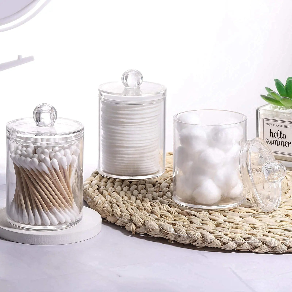 Four clear glass storage jars with lids filled with white cotton swabs arranged on a woven mat.
