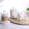 Four clear glass storage jars with lids filled with white cotton swabs arranged on a woven mat.