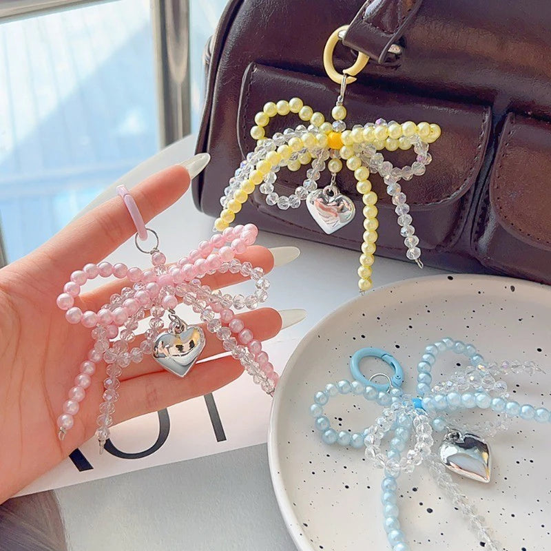 A hand holds a pink pearl bow keychain with a silver heart charm next to a dark brown leather bag.