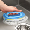 A hand is holding a blue sponge cleaning brush with an ergonomic handle above a metal strainer.