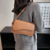 A woman wearing a grey sweater holds a tan vintage leather crossbody bag with a flap closure.