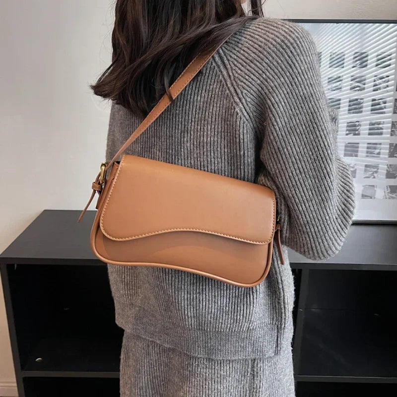 A woman wearing a grey sweater holds a tan vintage leather crossbody bag with a flap closure.
