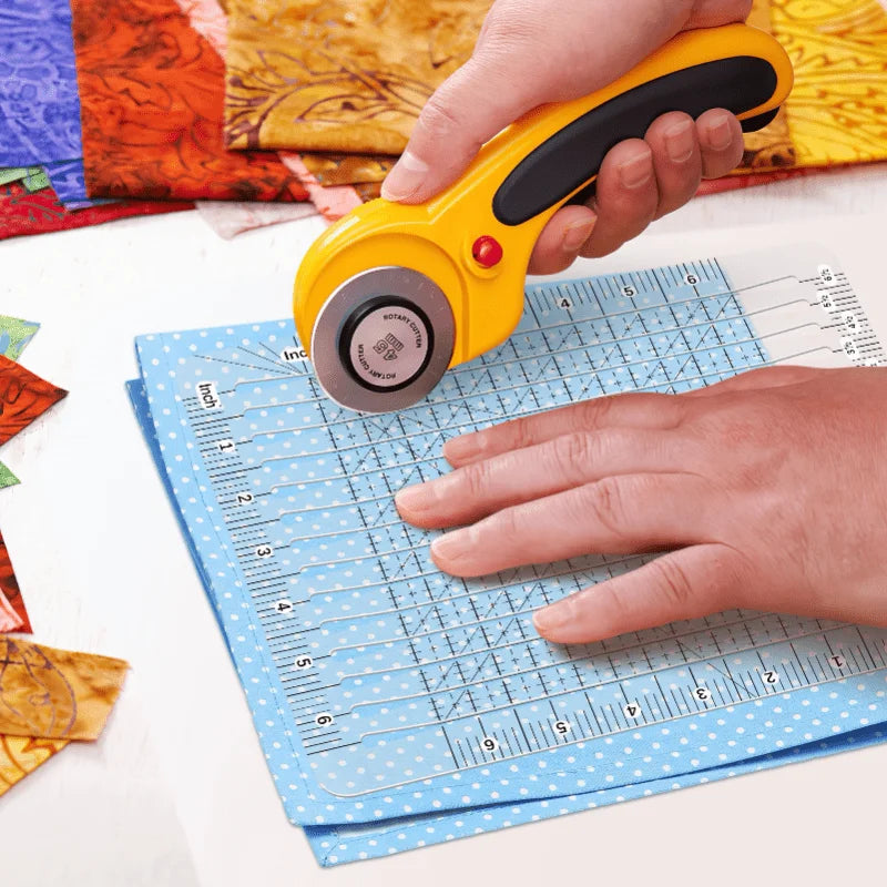 A hand holds a yellow acrylic quilting ruler with a dual scale grid and seam guide on a blue paper.