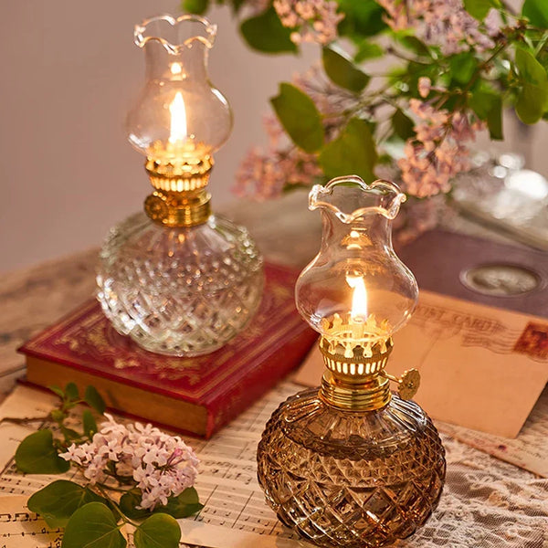 Lampe à huile vintage rustique en verre et laiton sur une surface en bois, à côté d'un livre et d'éléments floraux.