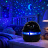 A black projector night light with blue LED lights is displayed on a bedside table under a starry sky theme.