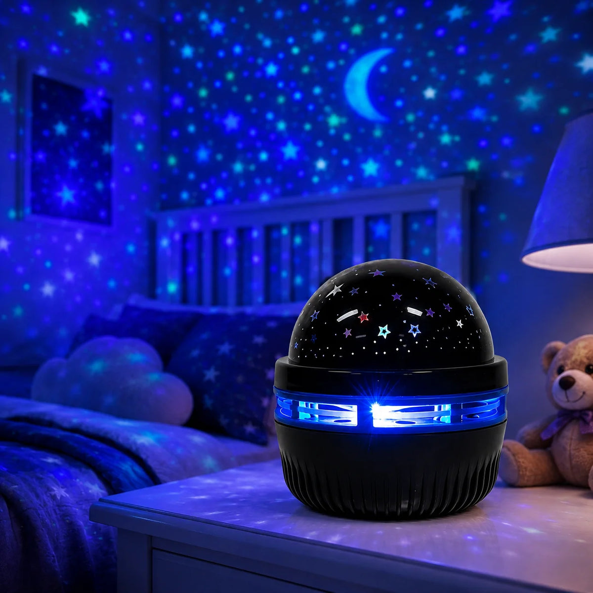 A black projector night light with blue LED lights is displayed on a bedside table under a starry sky theme.