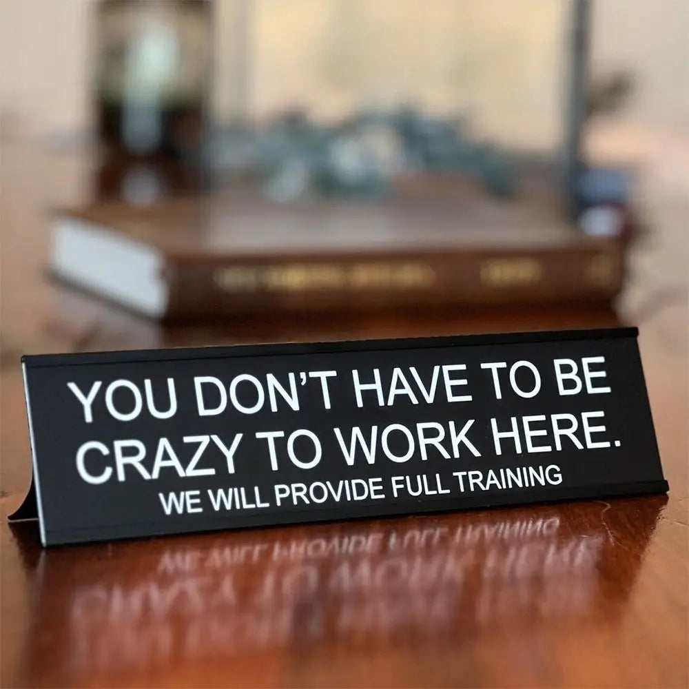 A black desk sign with white text that reads You don't have to be crazy to work here we will provide full training