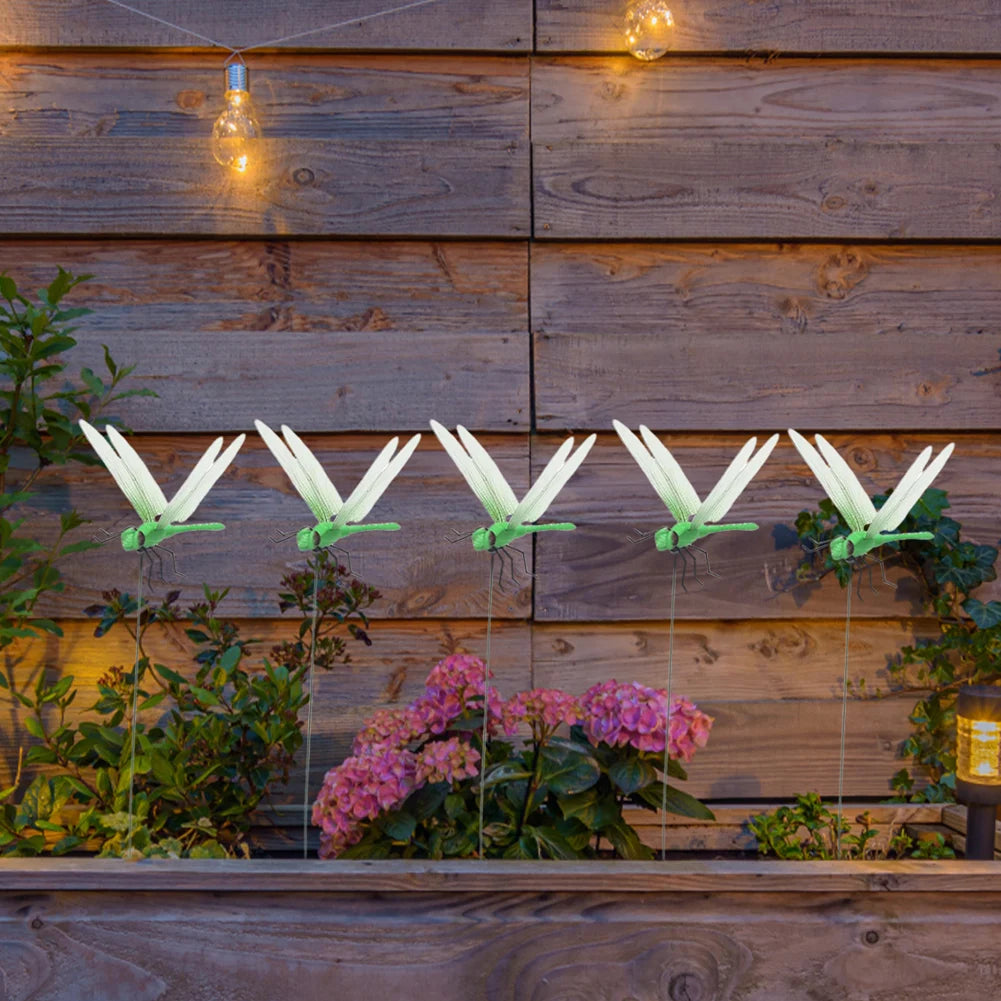 Lifelike dragonfly clips hanging from a wooden panel with plants and string lights.