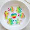 A white bowl contains several iridescent, colorful resin charms shaped like fish and sea creatures for crafting.