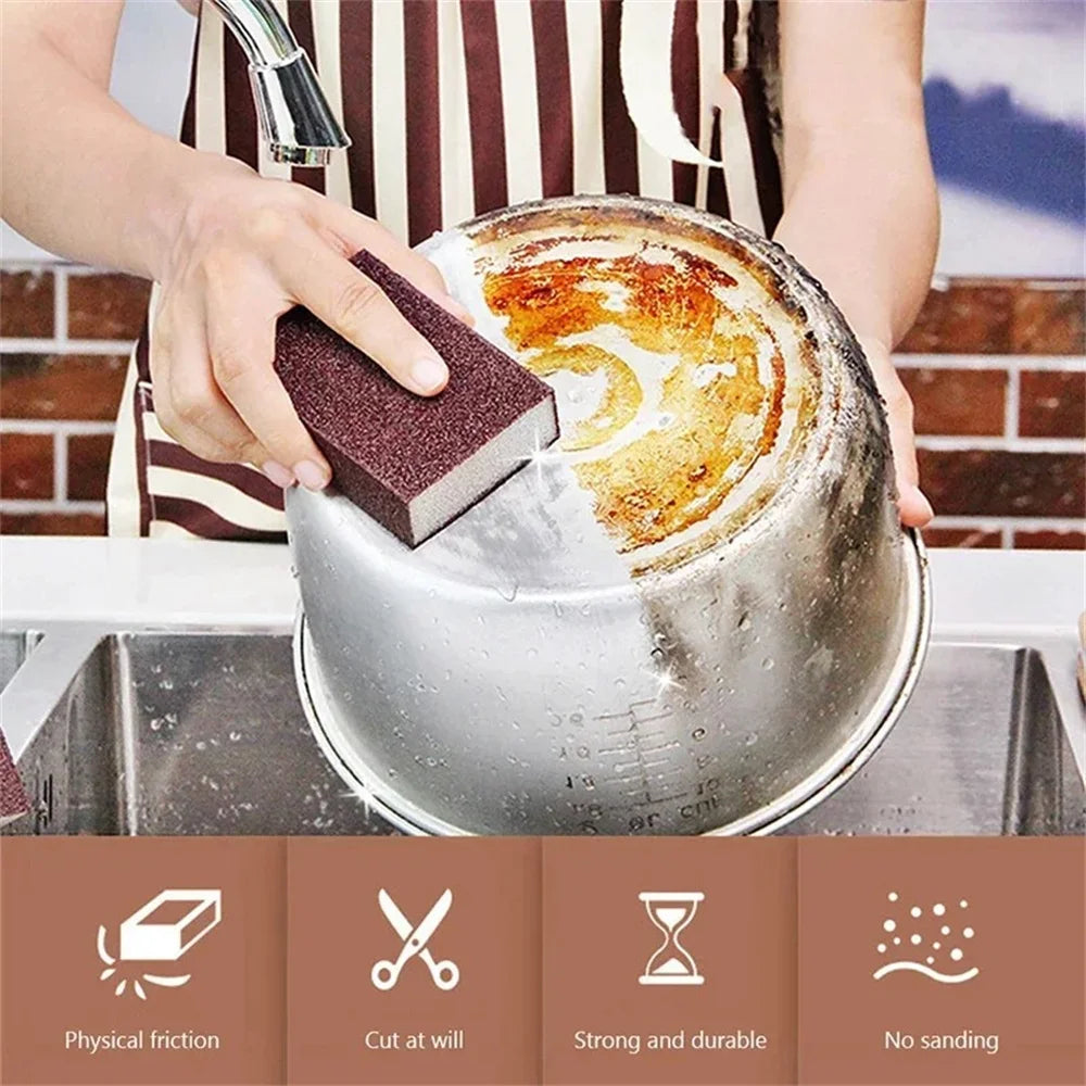 A hand is using a brown cleaning sponge to scrub a stainless steel pot in a kitchen setting.