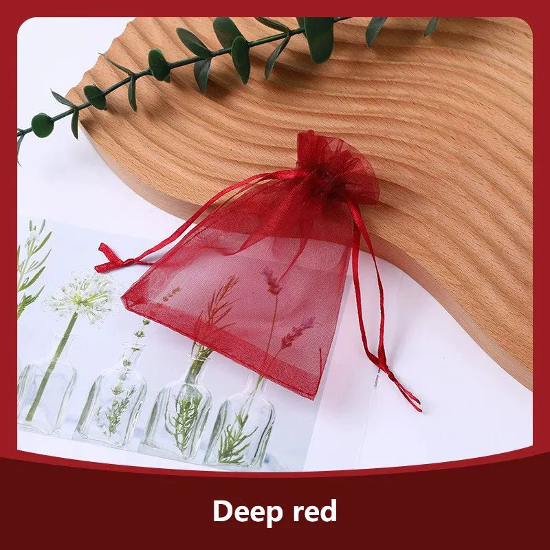 A deep red organza drawstring gift bag tied with a ribbon resting on a wooden surface next to small glass vials.