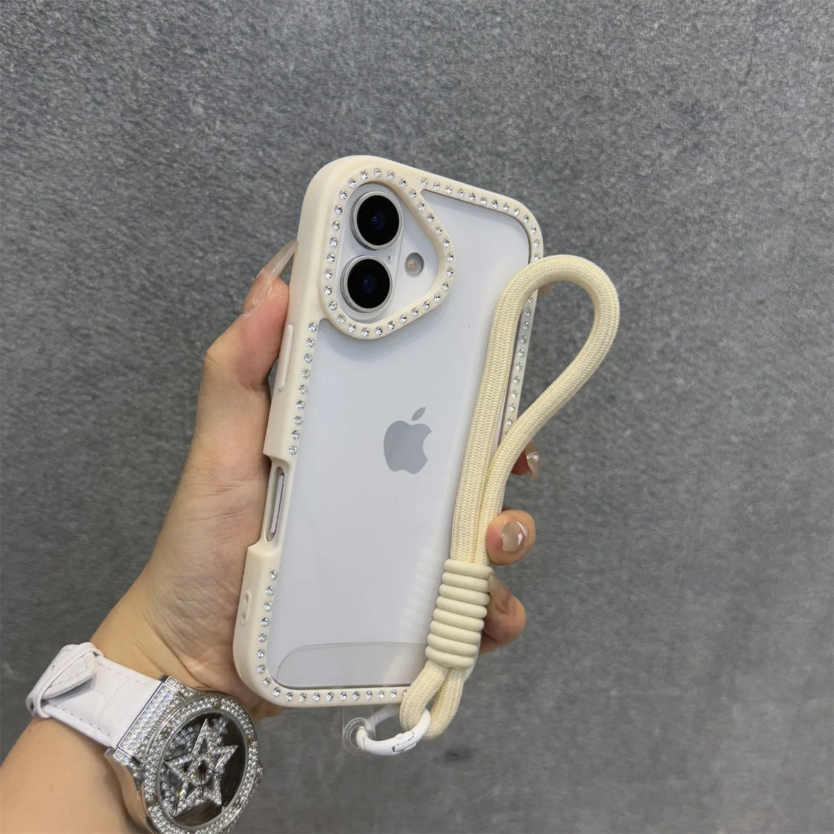 Una funda blanca para iPhone con diseño transparente de diamante y una correa beige sostenida en una mano.