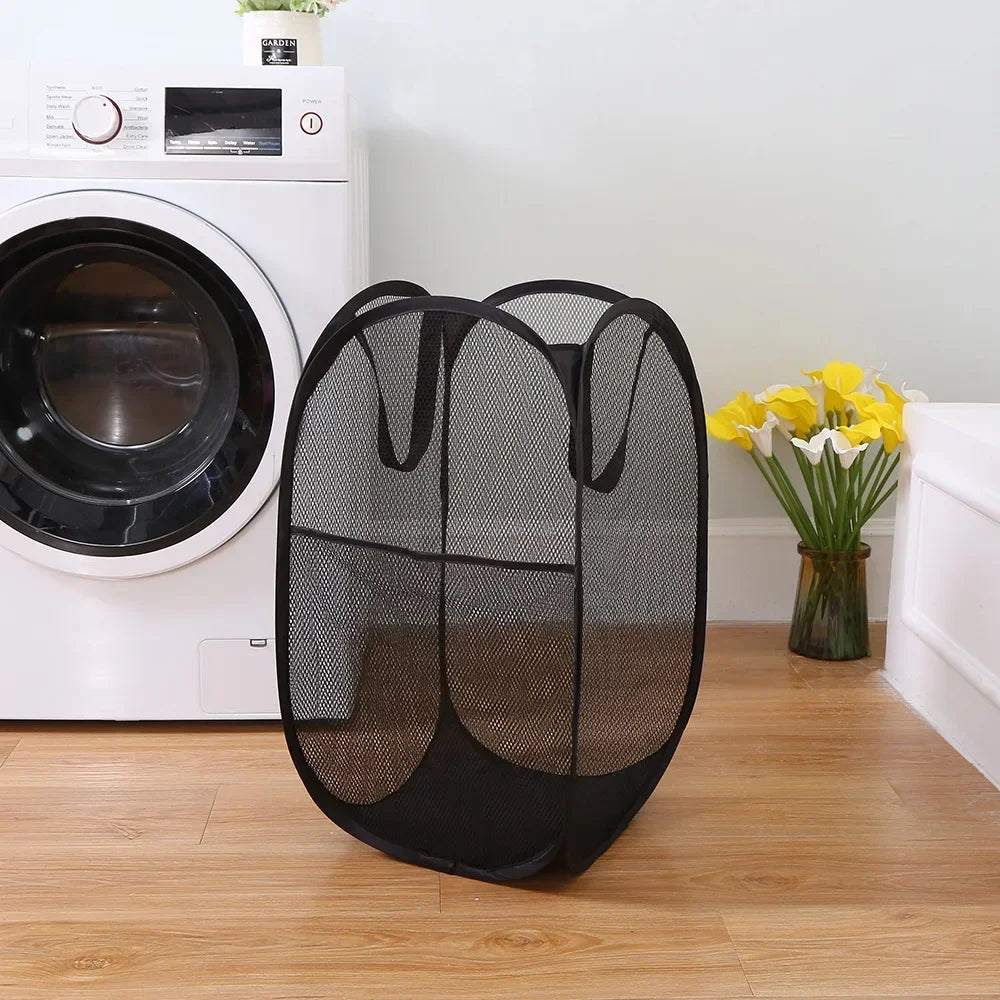 A black foldable laundry hamper with a mesh design sits on a wooden floor next to a white washing machine.