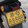 A yellow license plate with black characters is mounted on a dark vehicle, showing the plate and mounting hardware.