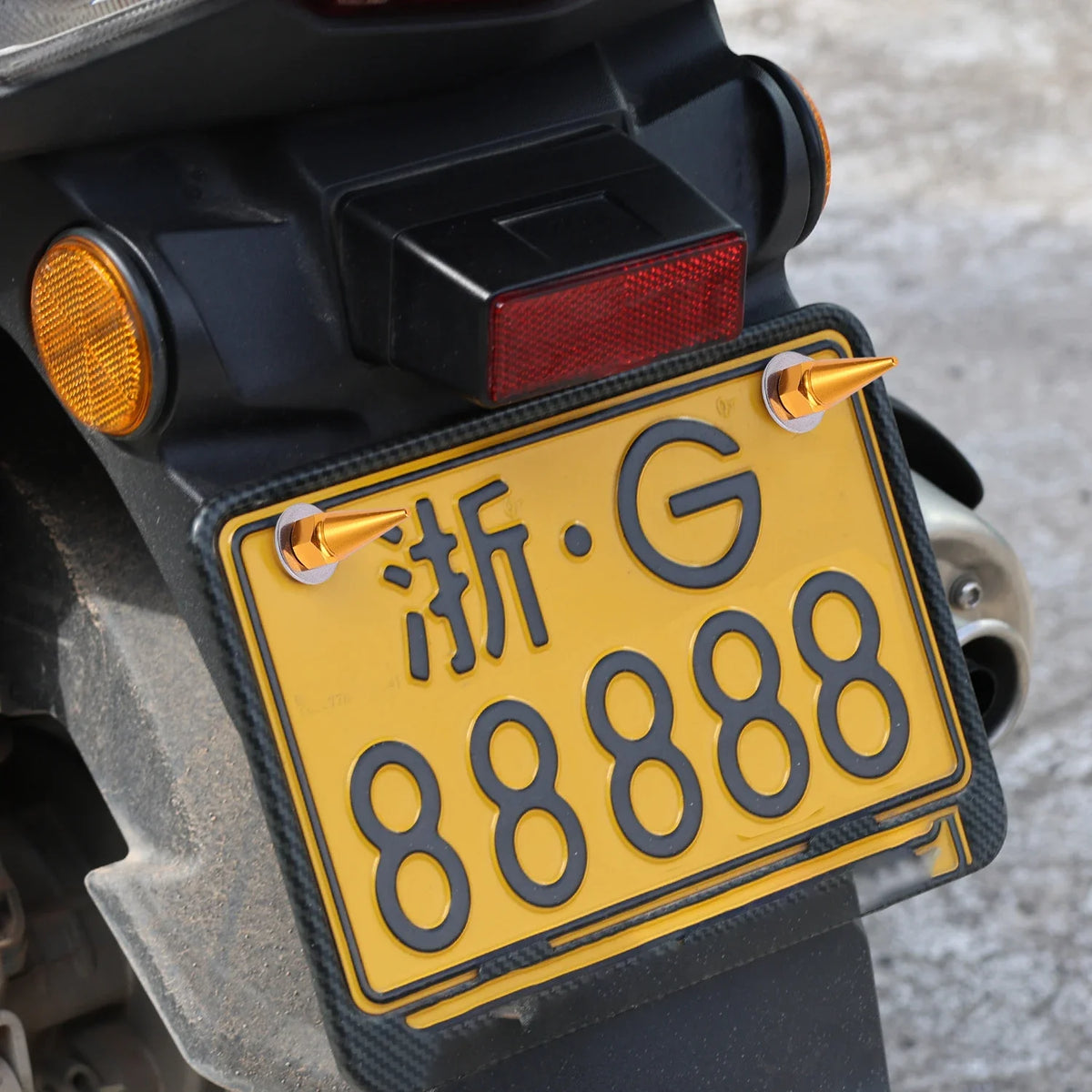 A yellow license plate with black characters is mounted on a dark vehicle, showing the plate and mounting hardware.