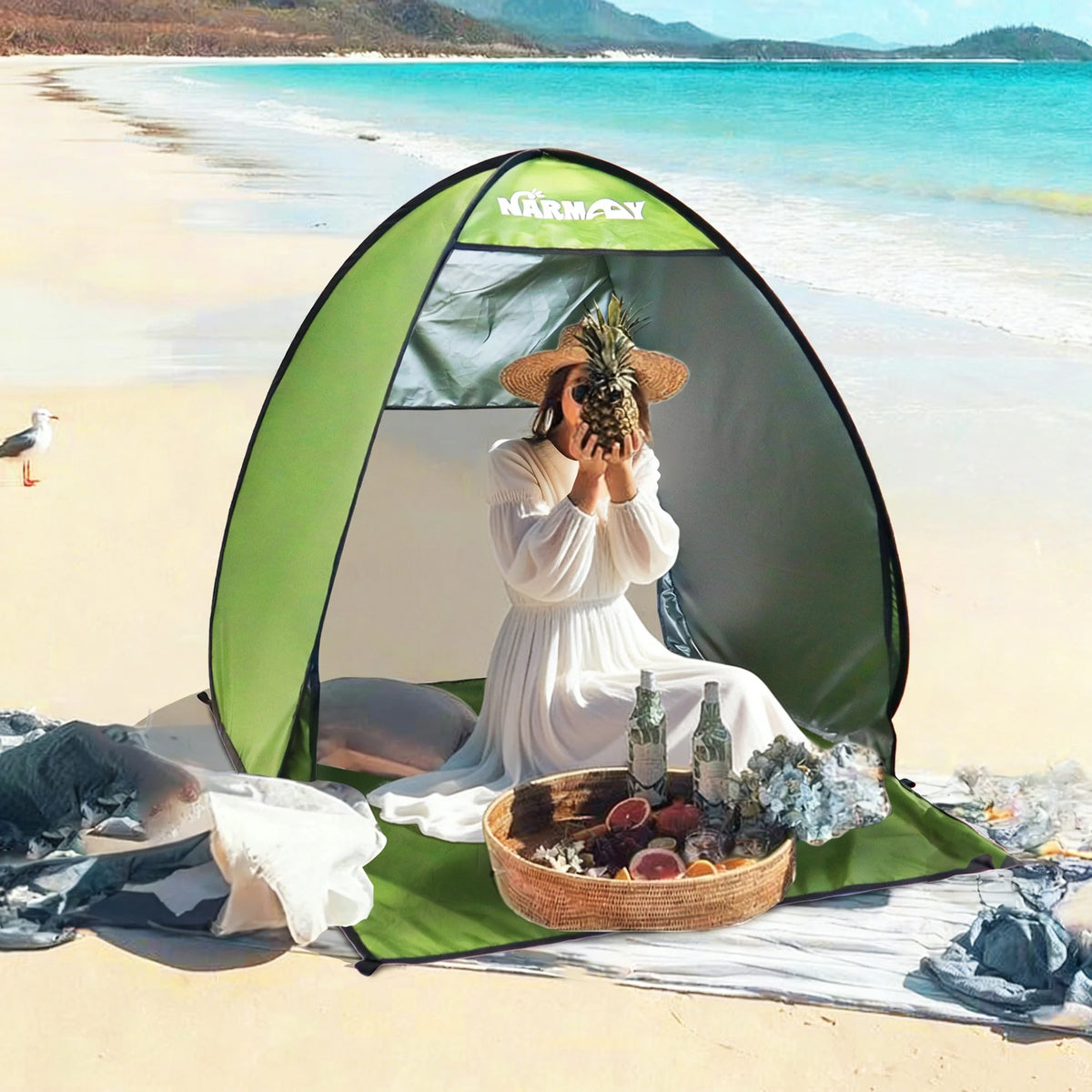 A white dress model sits inside a green pop-up beach tent on a sandy beach with tropical foliage and sea in the background.