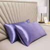 A stack of silky smooth purple satin pillowcases resting on a light surface next to a lamp.