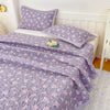 Purple floral quilted bedspread with pink flowers draped over a white bed in a bedroom setting.
