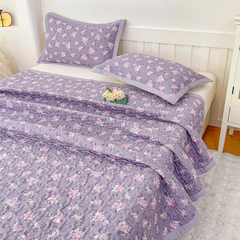 Purple floral quilted bedspread with pink flowers draped over a white bed in a bedroom setting.