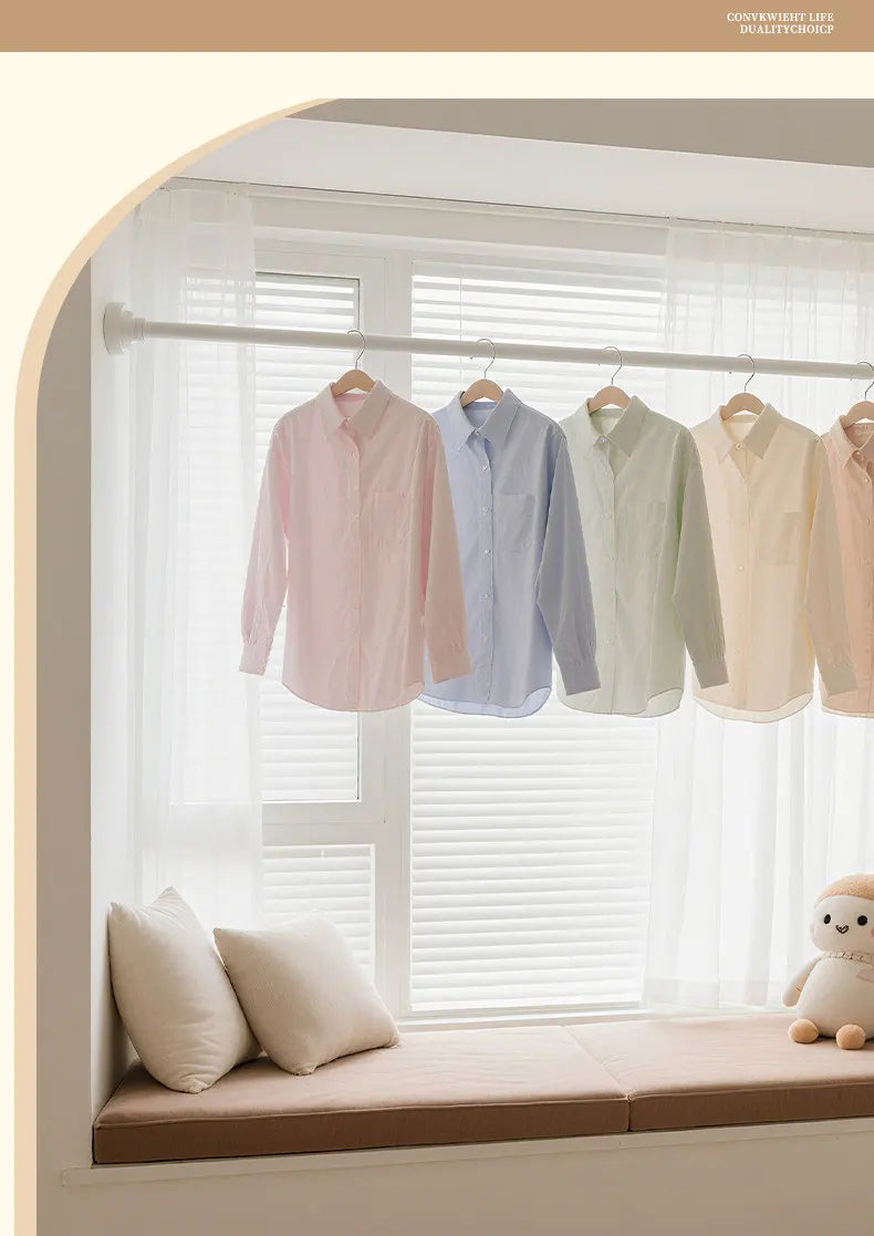 A collection of light-colored shirts hanging on a shower curtain rod in a bright window setting.