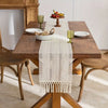 A wooden dining table set for a meal with a beige tasseled table runner and fruit