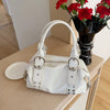A white soft PU leather shoulder bag with silver rivet accents resting on a light wooden surface next to a vase of red flower