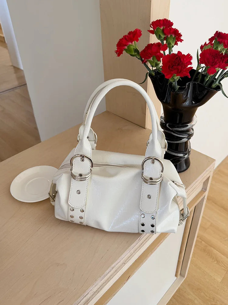 A white soft PU leather shoulder bag with silver rivet accents resting on a light wooden surface next to a vase of red flower
