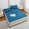 A teal waterproof mattress protector with elastic straps is displayed on a bed, next to a white chair and a bedside table.