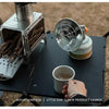 A portable butane gas stove is set up on a dark surface outdoors, with wood fuel and a metal strainer visible.