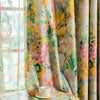 A close-up of a light floral plant pattern curtain panel in shades of yellow, pink, and green hanging next to a window.