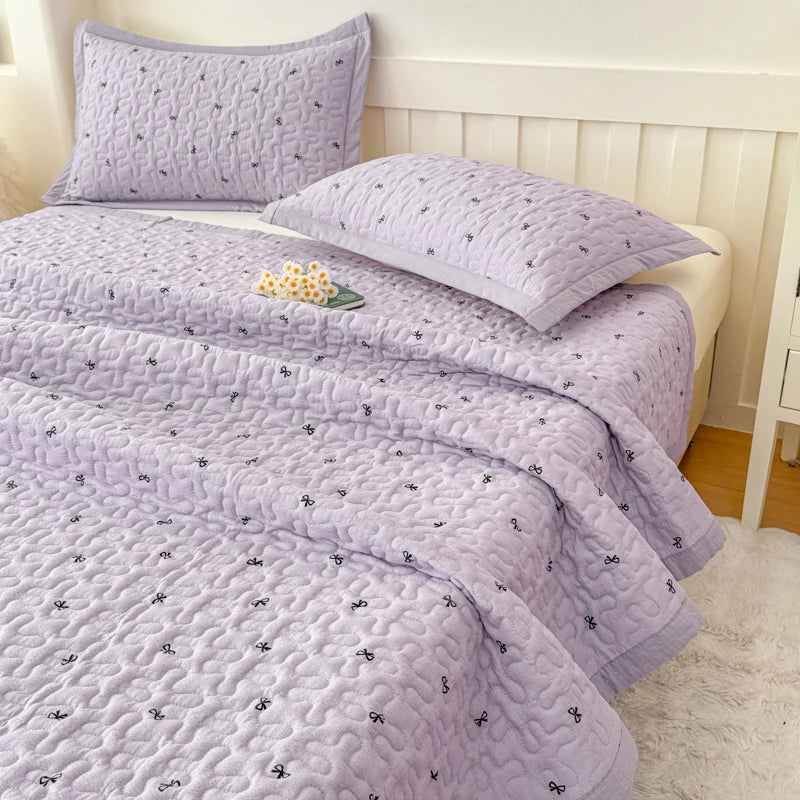 A purple quilted bedspread with a subtle floral pattern is displayed on a bed, featuring a white headboard and light-colored 