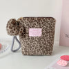 A brown leopard print cosmetic bag with a fluffy pom-pom and gray strap is displayed on a white surface.
