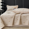 A beige quilted bedspread with a light gingham pattern featuring small gold bear embroidery, displayed on a dark headboard.