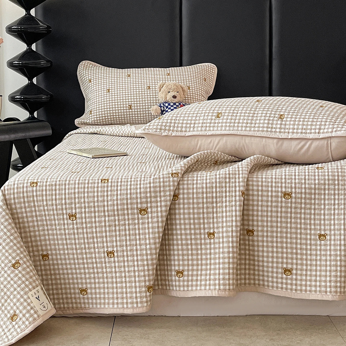 A beige quilted bedspread with a light gingham pattern featuring small gold bear embroidery, displayed on a dark headboard.