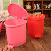 A pink strawberry-shaped mini trash can with a lid sits on a wooden surface next to a red one.