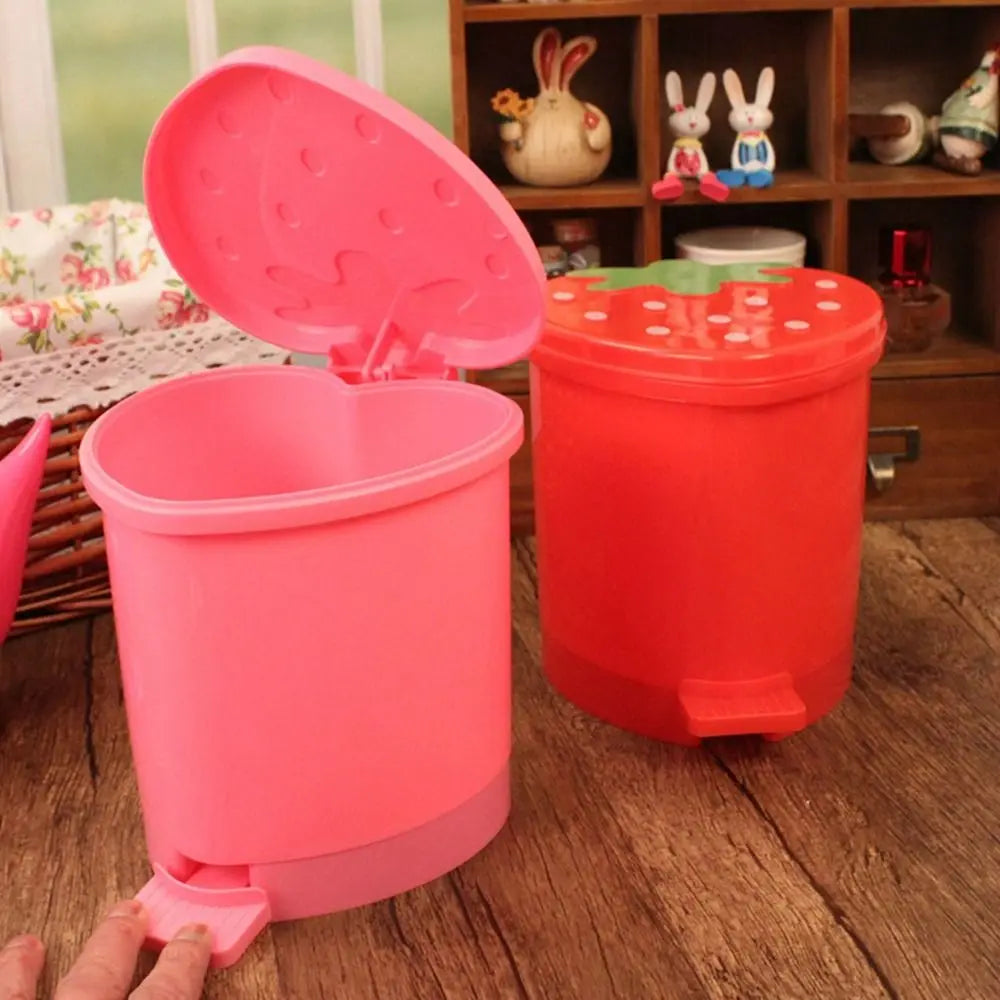 A pink strawberry-shaped mini trash can with a lid sits on a wooden surface next to a red one.