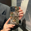 A hand holds a phone with a forest camouflage silicone case featuring tree branches and leaves.