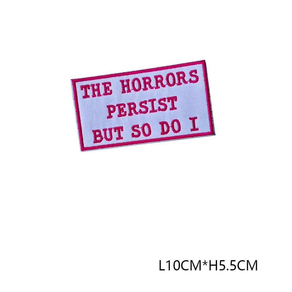 A pink embroidered letter patch with black text that reads The Horrors Persist But So Do I
