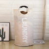A large, beige foldable laundry bag with a padded black handle is displayed against a white brick wall.