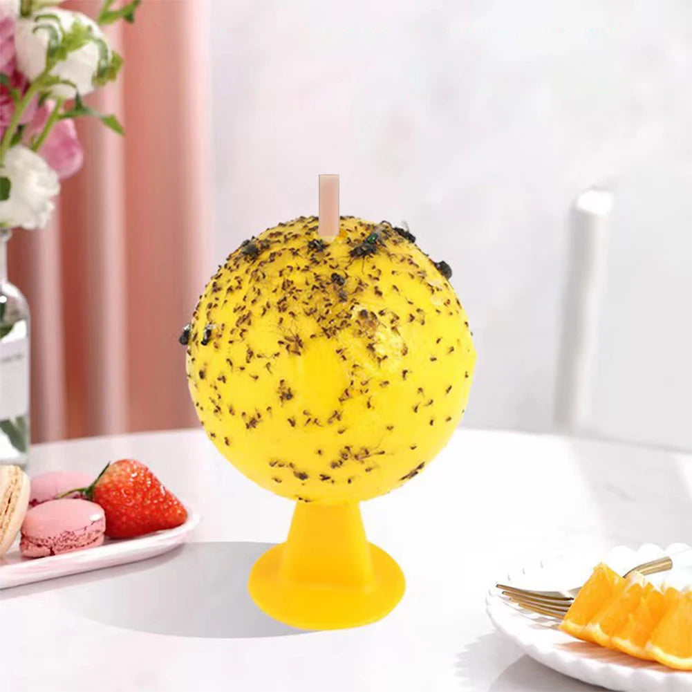 A yellow sticky fruit fly trap ball with black specks is displayed on a white surface.