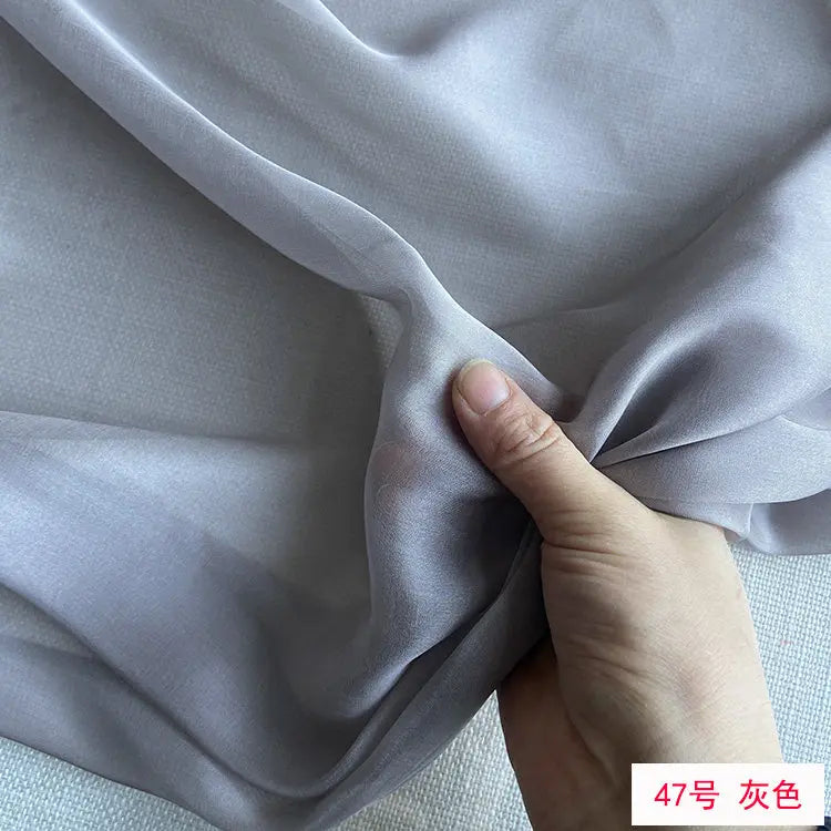 A hand is gently folding a piece of light gray mulberry silk chiffon fabric.