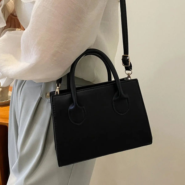 Bolso cruz negro de cuero PU clásico llevado por una persona con camisa blanca y pantalones gris claro.
