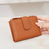 A tan leather compact zippered wallet with a quilted pattern is held in a hand on a light surface.