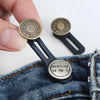 A hand holds an adjustable metal jeans button extender with a round gold-toned top and black base against denim fabric.