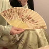A person holds a light beige bamboo and silk folding fan with floral designs.