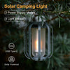A gray solar camping light lantern with a flickering flame inside, hanging from a branch in an outdoor setting.