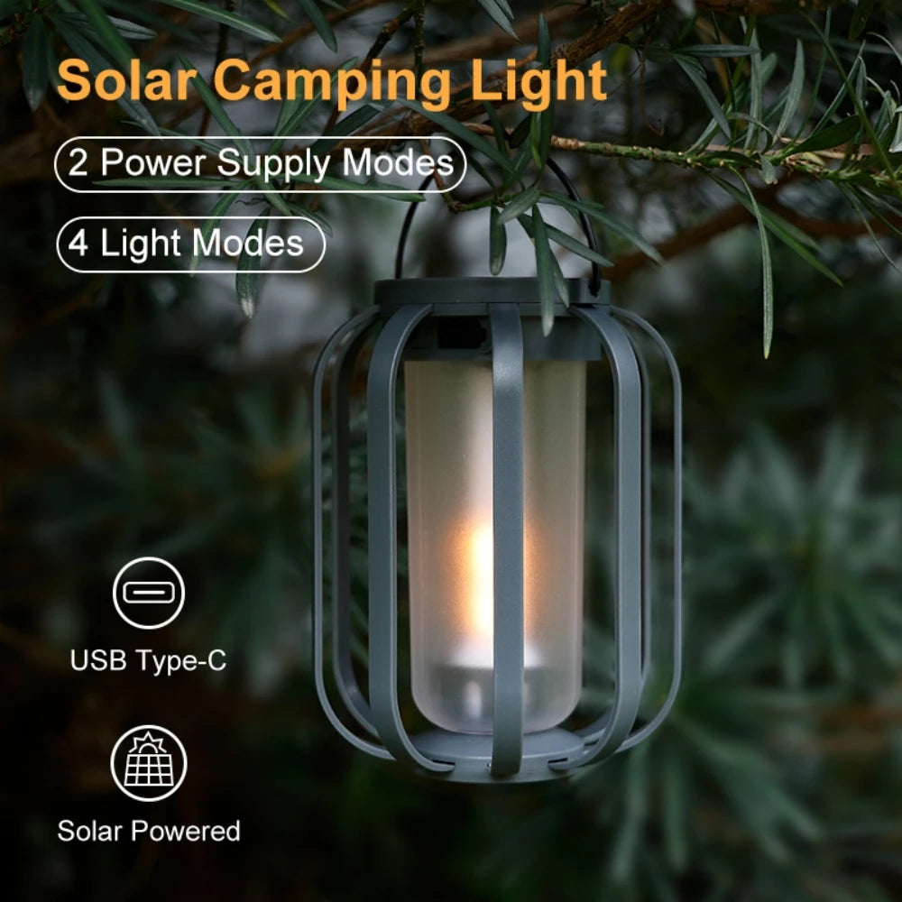 A gray solar camping light lantern with a flickering flame inside, hanging from a branch in an outdoor setting.